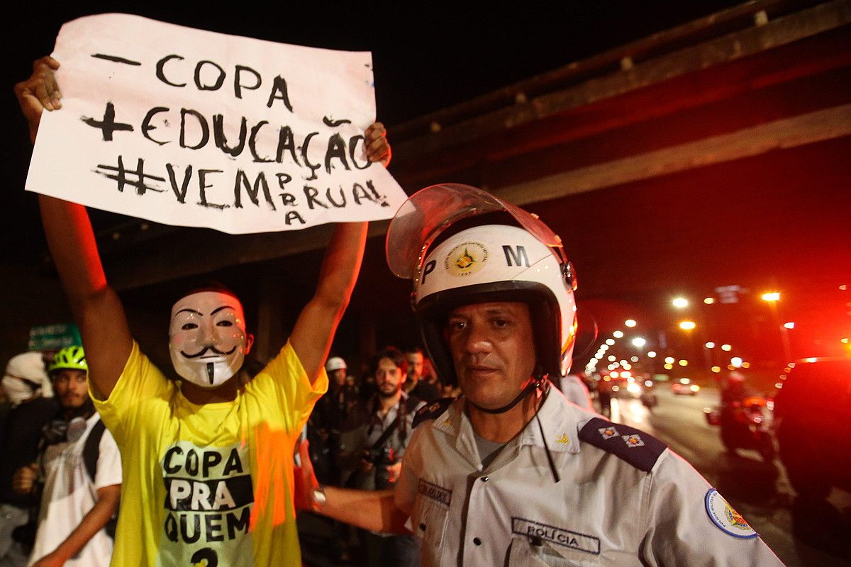 Maskara batekin aurpegia estalia duen manifestari bat Kopa gutxiago, hezkuntza gehiago; etor zaitez kalera dioen afixarekin, ondoan polizia bat duela, Brasilian egindako protesta batean. FERNANDO BIZERRA JR. / EFE.