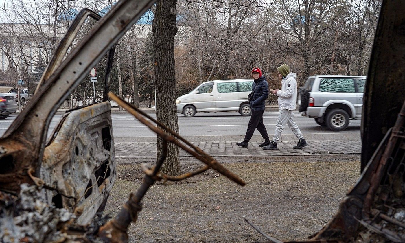 Bi pertsona, atzo, Almatyn, protestetan kiskalitako ibilgailu baten aurretik pasatzen. EFE.