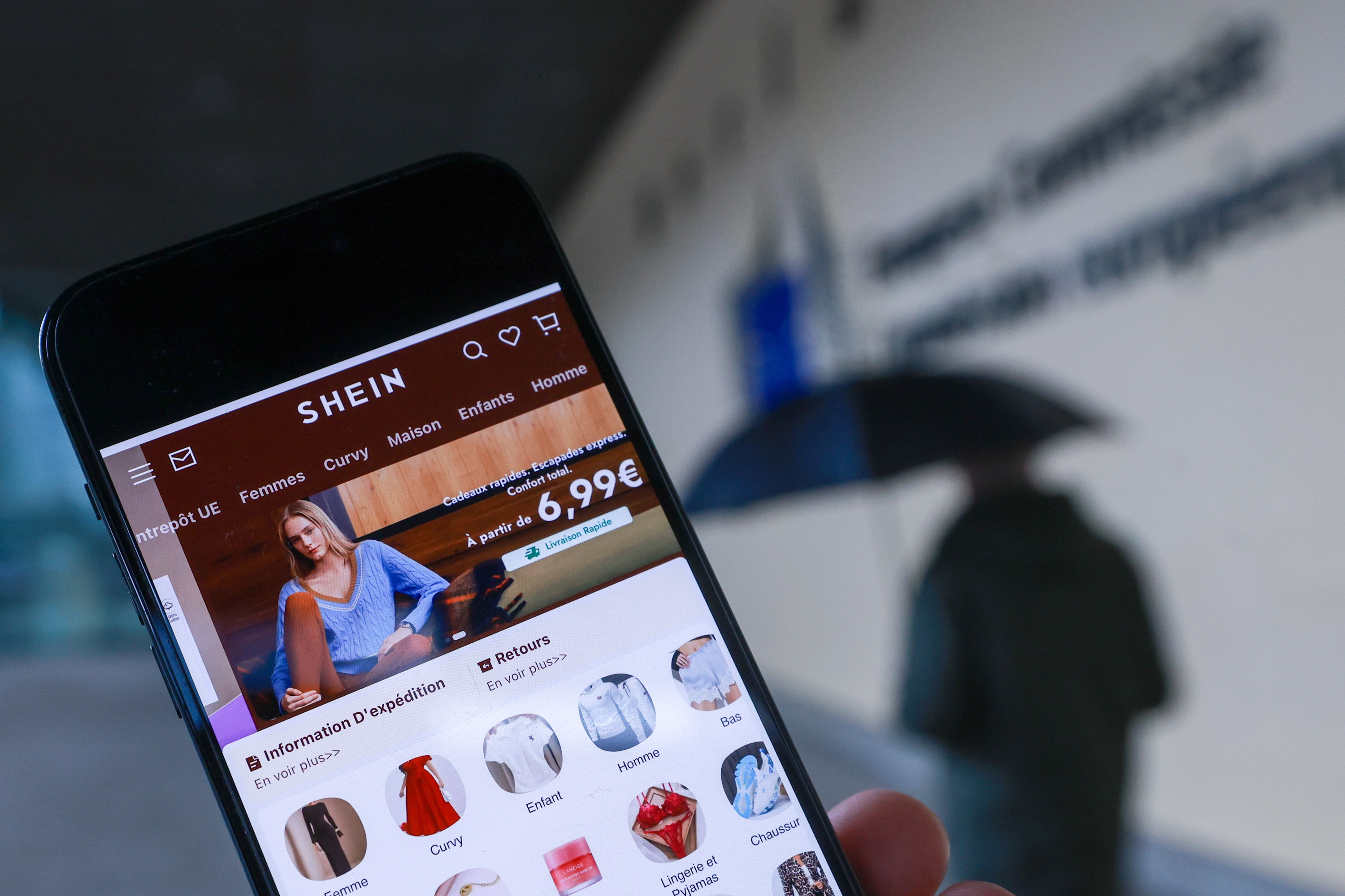 Sheinen webgunea eskuko telefono batean, eta Europako Batzordearen egoitza atzean. OLIVIER HOSLET / EFE